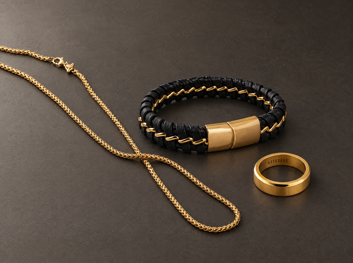 Akitsune Franco gold necklace, Adeptus black leather and gold bracelet and Purus gold ring arranged on a charcoal surface.
