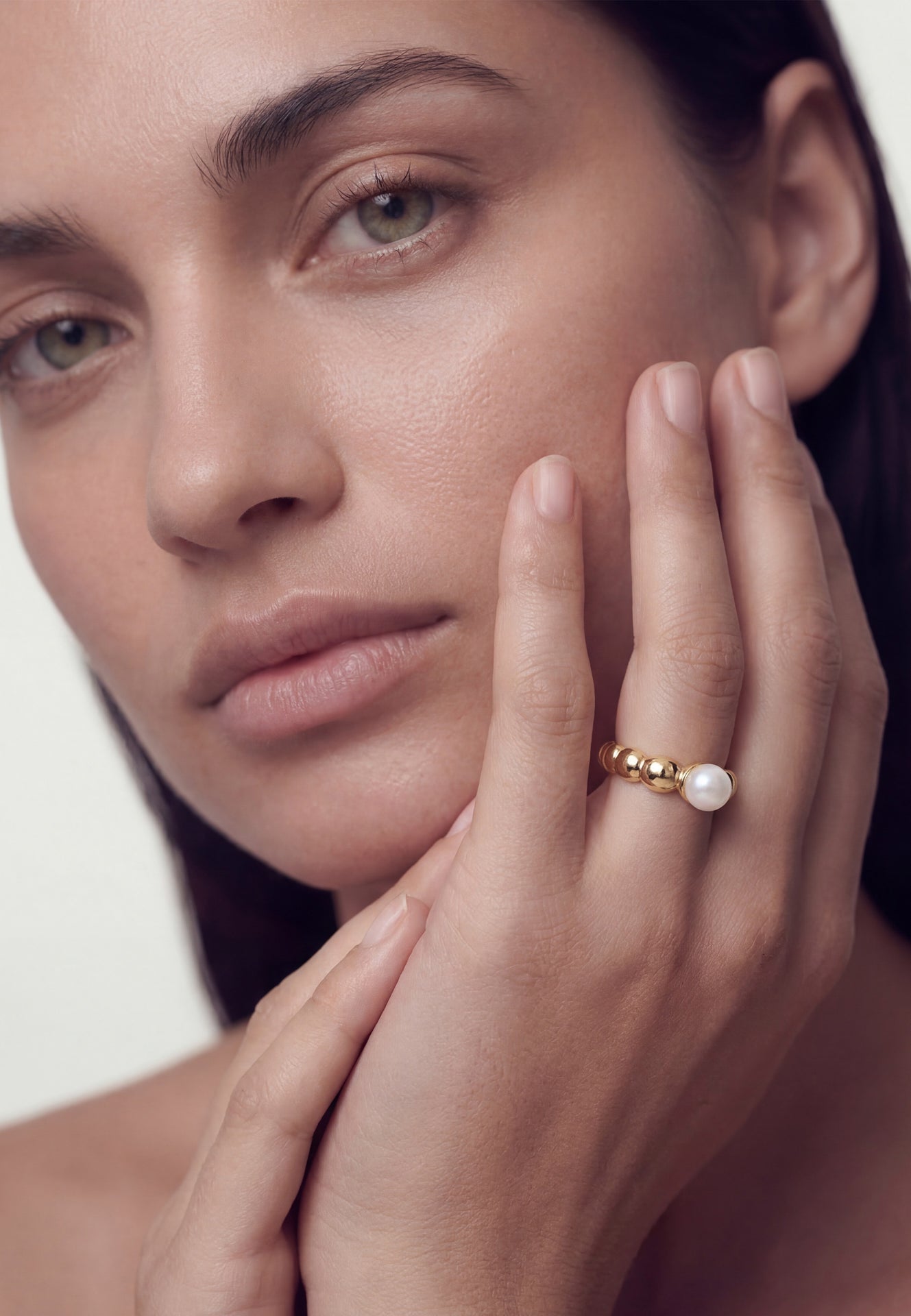 Close-up of a woman's face with a pearl ring on her finger against a neutral background
