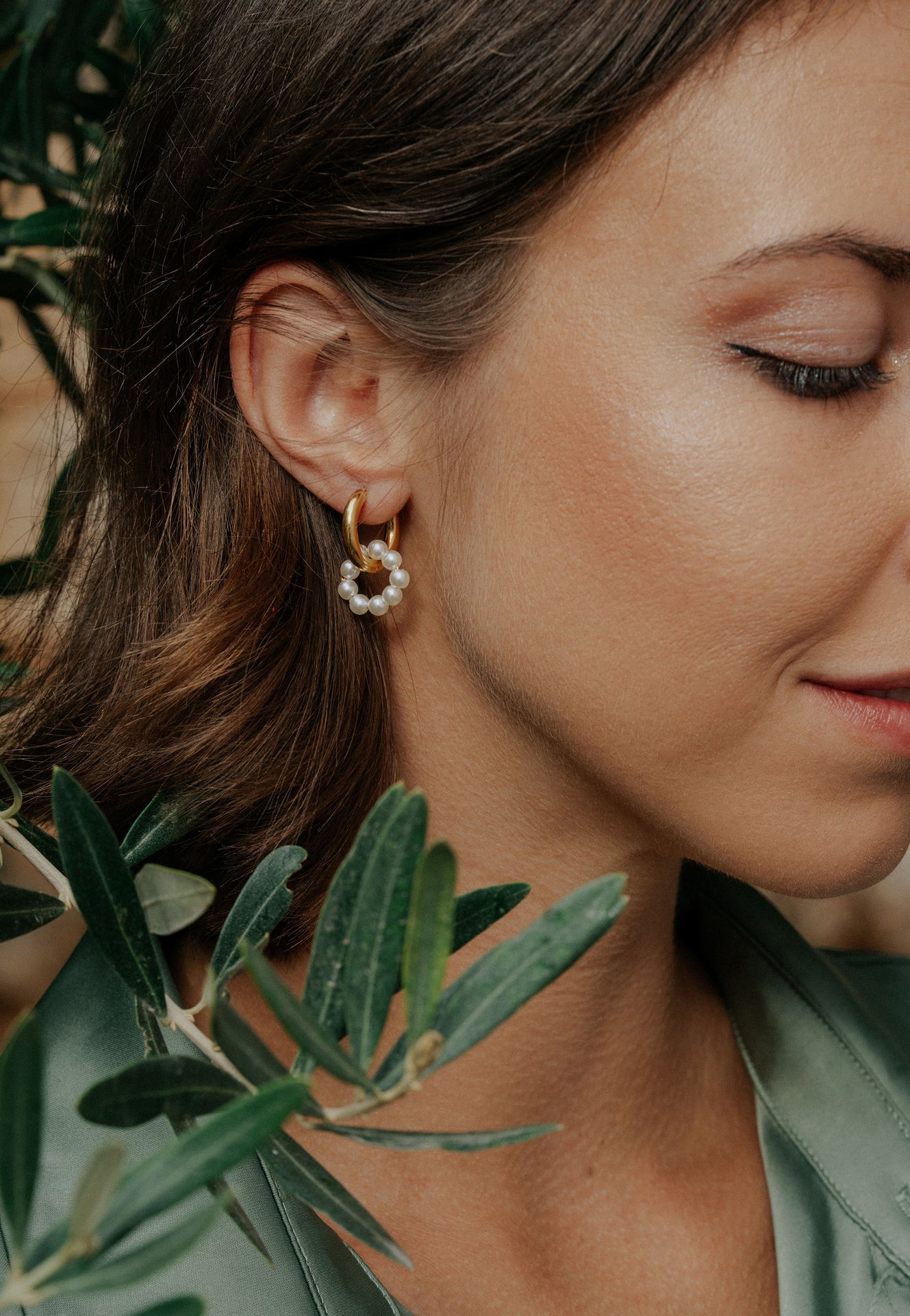 Close-up of a woman wearing gold hoop earrings with small pearls, surrounded by green leaves.
