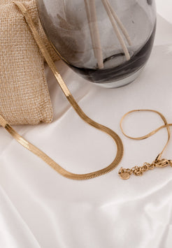 Gold necklace and bracelet on a white surface with a marble-patterned vase in the background.