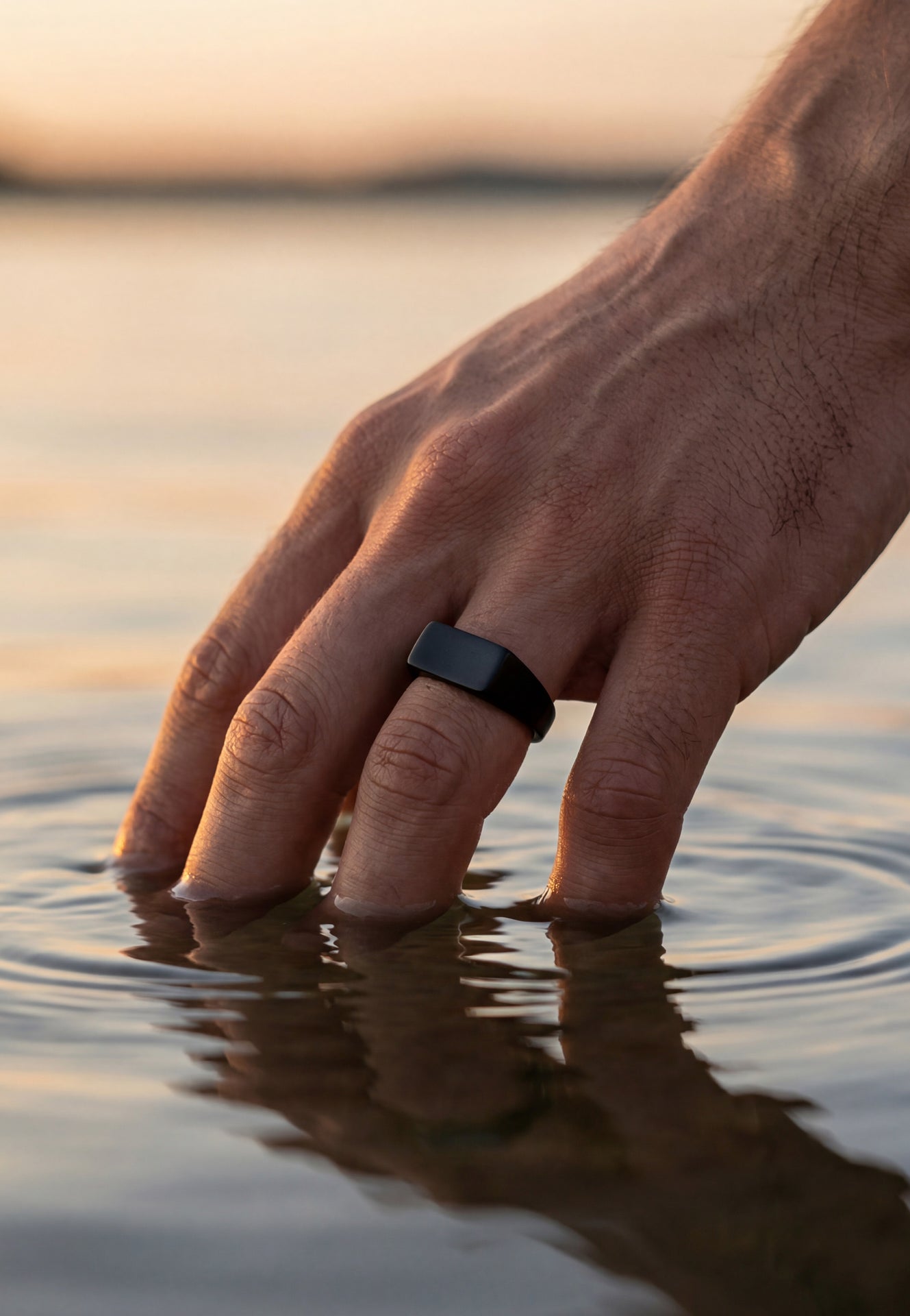 Hand with a black ring touching water at sunset