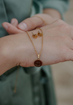 Gold necklace with smiley face pendant on a hand, with earrings in the background.