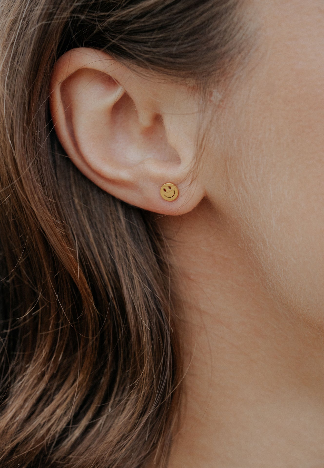 Close-up of an ear wearing a gold smiley face earring with blurred hair and skin in the background.