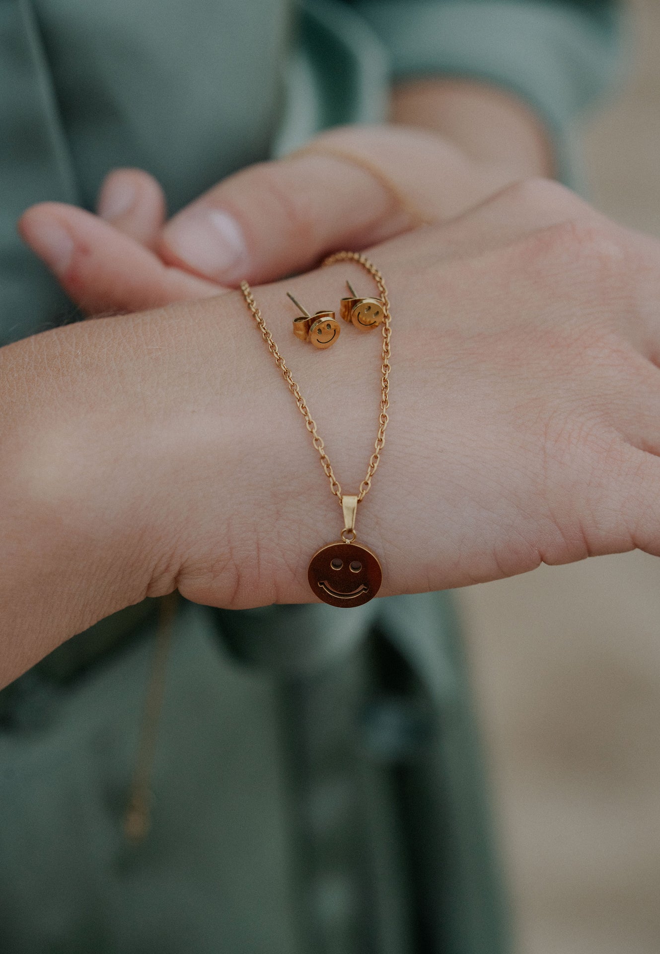 Gold smiley face necklace and earrings on a wrist with a blurred background