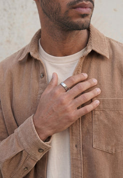 Male model wearing a minimalist silver band ring on finger with a beige corduroy overshirt, showcasing a modern men's jewelry design.