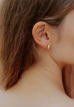Close-up of an ear wearing a gold teardrop earring with blurred hair and skin in the background