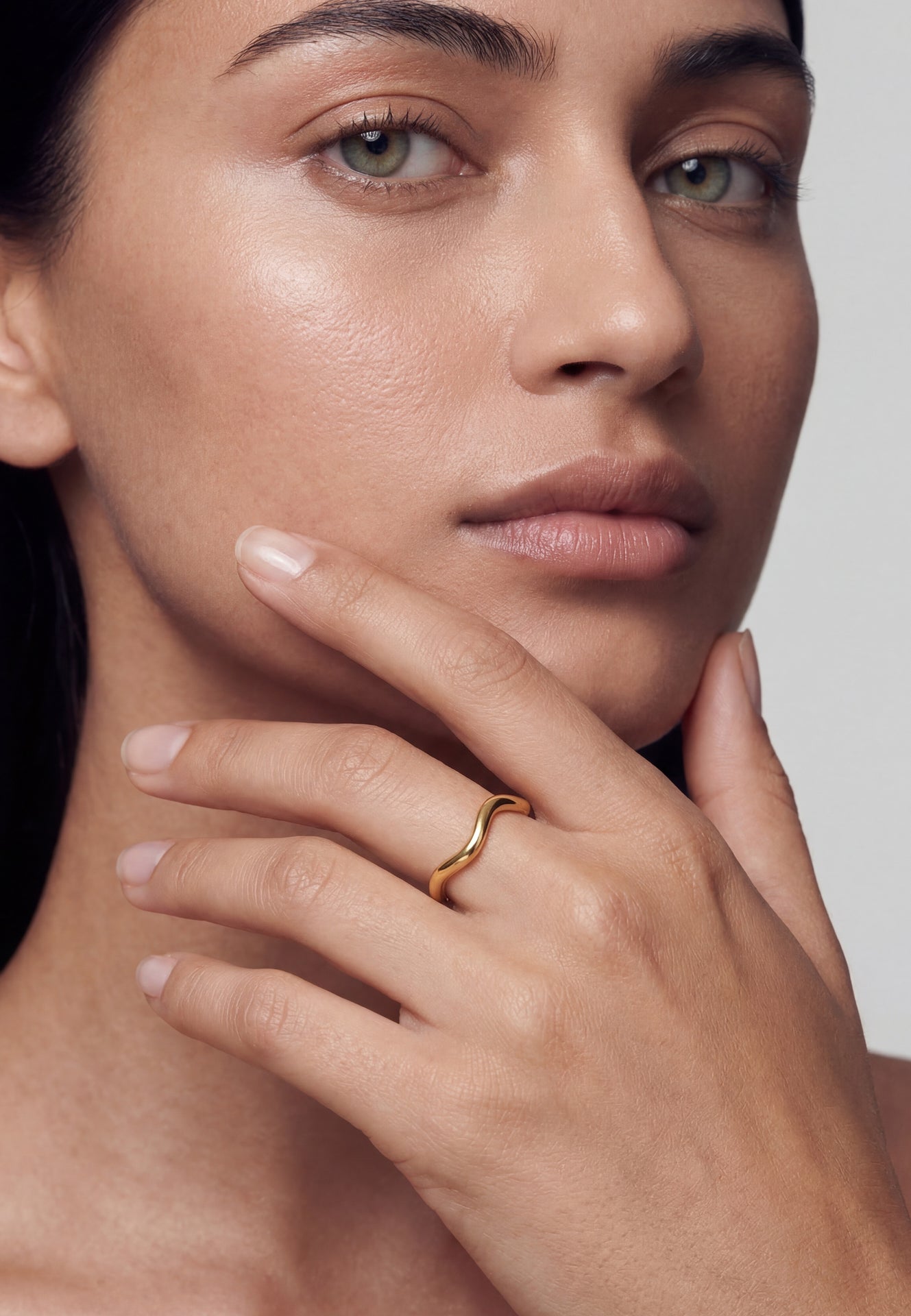 Close-up of a woman's face with a gold ring on her finger, against a neutral background