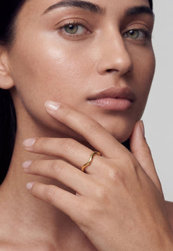 Close-up of a woman's face with a gold ring on her finger, against a neutral background