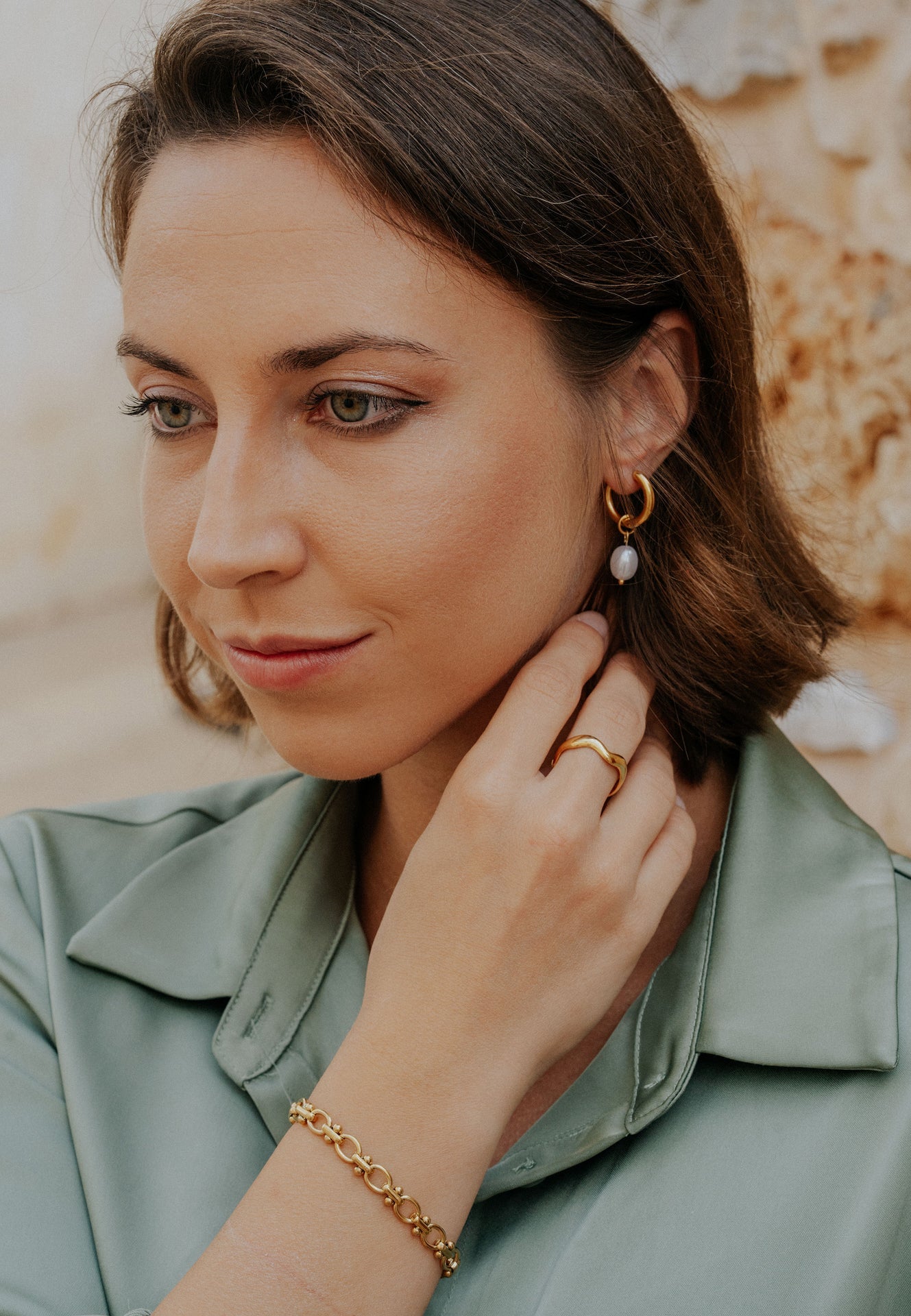 Woman adjusting an earring with a gold bracelet, wearing a light green top.
