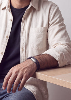 A close-up of a man's arm resting on a light wood table, wearing a dark braided bracelet with a silver-toned rectangular metal link.