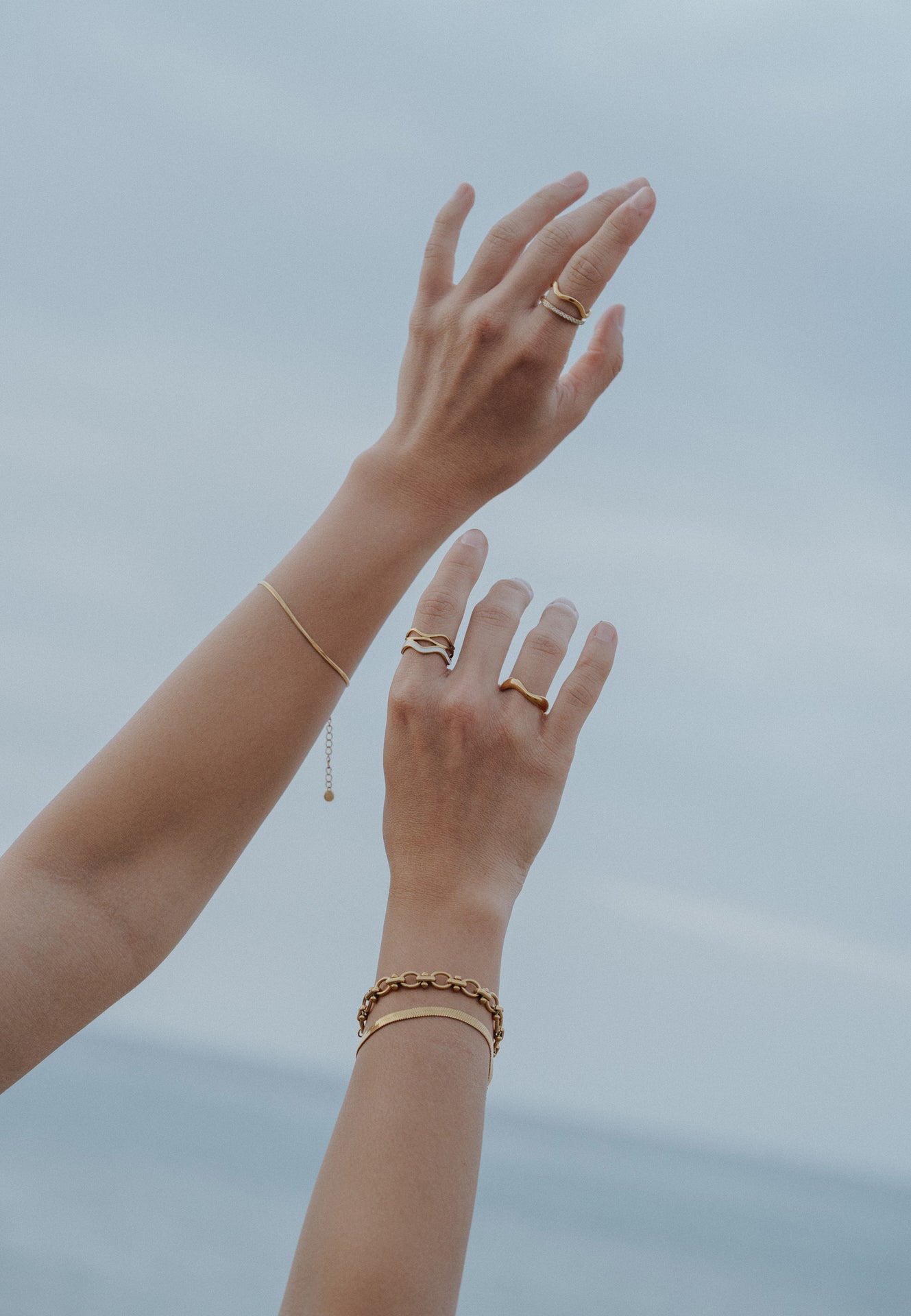 Two hands with gold jewelry reaching towards a blurred natural background